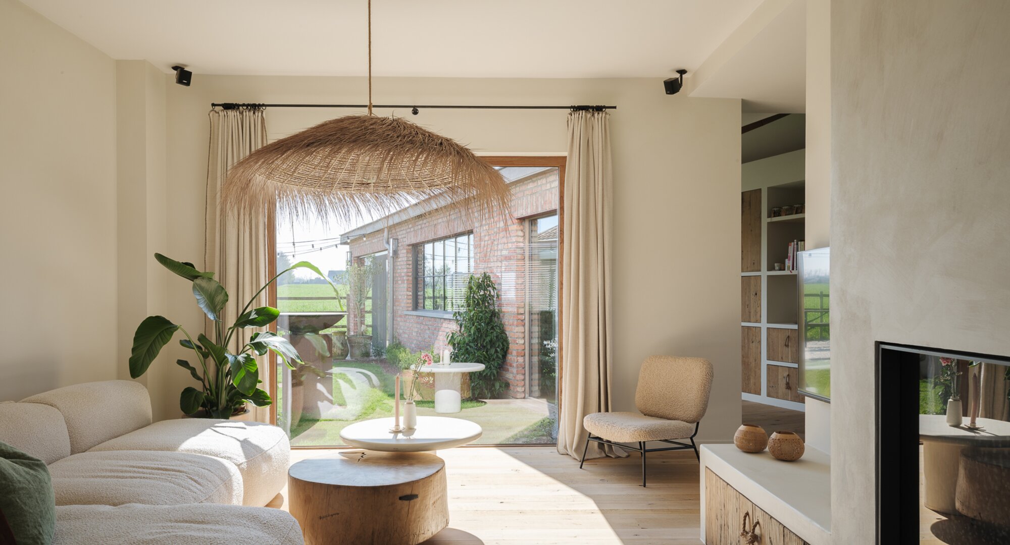 Lichte woonkamer met een beige hoekzetel, houten salontafels, een grote plant, een rieten hanglamp e