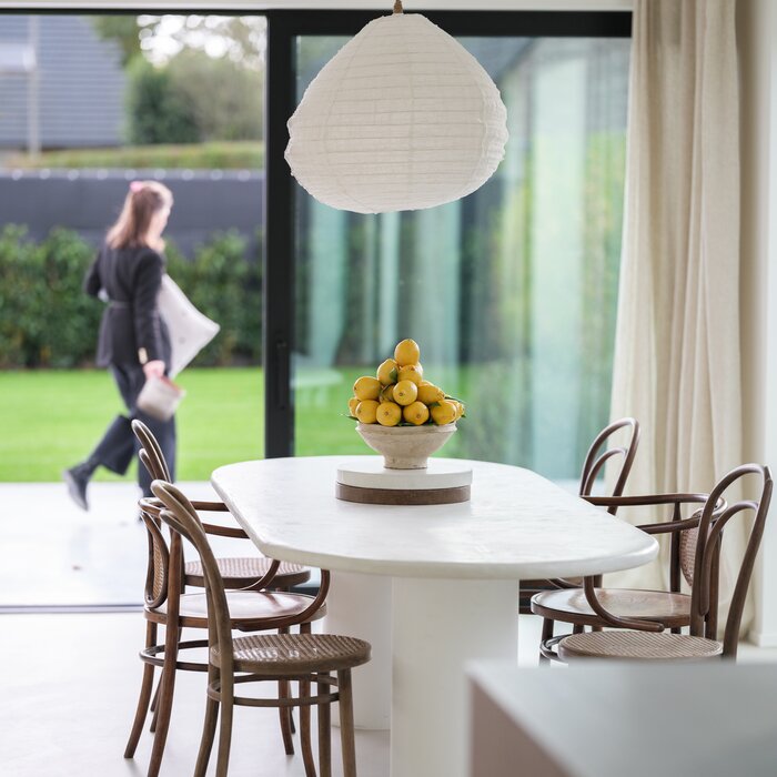 Eettafel met houten stoelen en een schaal citroenen in een moderne, lichte eetkamer met uitzicht op een tuin; op de achtergrond loopt een vrouw langs het raam.