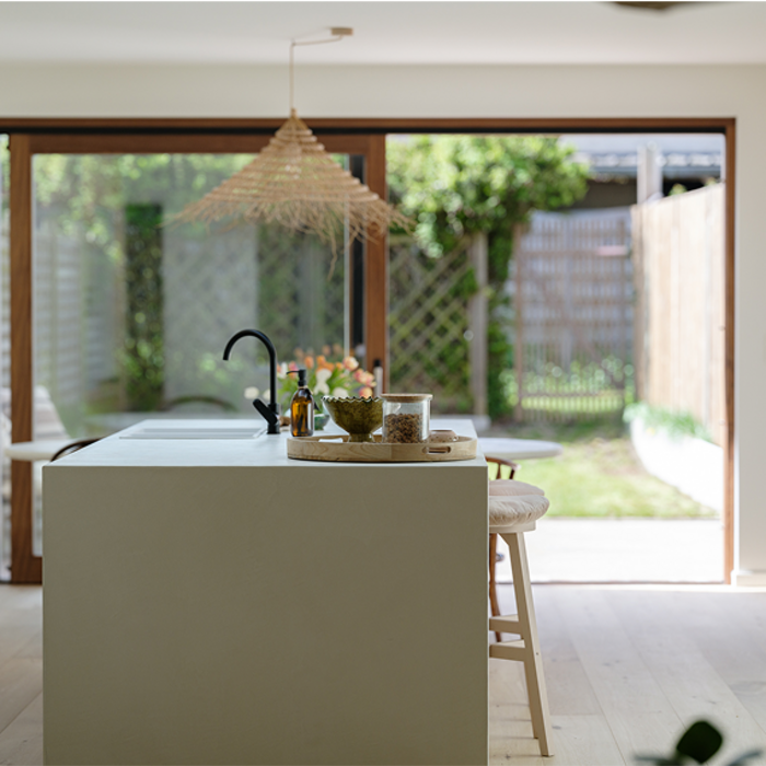 Moderne, minimalistische keuken met een lichtgekleurde kookeiland, zwarte kraan, houten kruk en glazen schuifdeuren die uitkijken op een zonnige tuin.